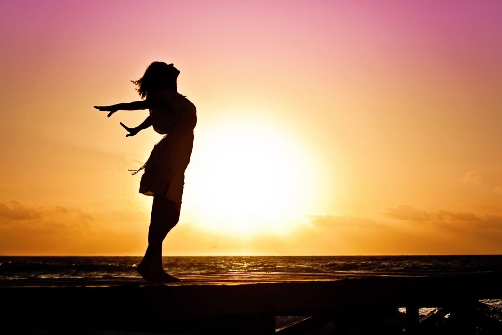 Terapias y Sesiones Silhouette of a woman at the beach with arms outstretched against a vibrant sunset backdrop.